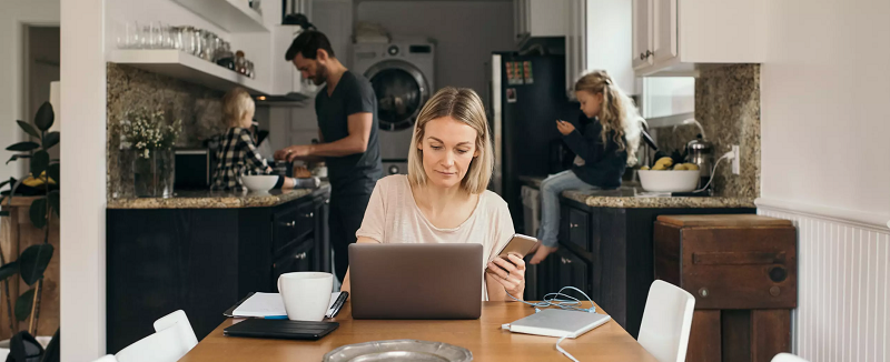 Woman is working remotely from home in the kitchen table with male and two kids in the background